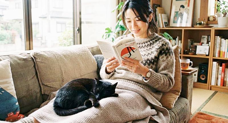 ソファで読書をする女性の膝の上で、丸くなって眠る黒猫